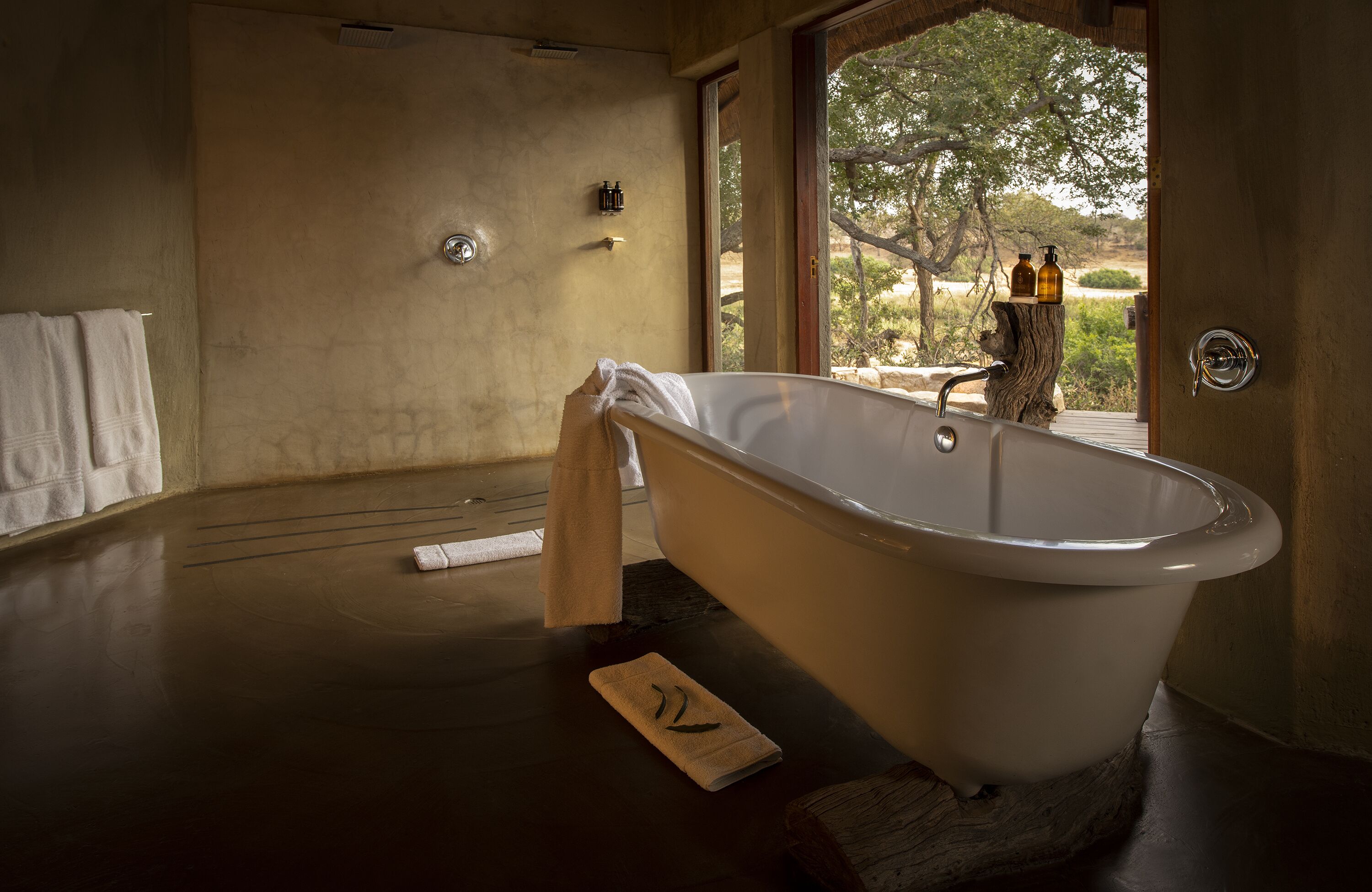 Ein geräumiges Badezimmer mit einer freistehenden Badewanne, Holzboden und Blick ins Freie durch ein großes Fenster.