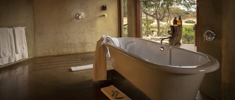 Ein geräumiges Badezimmer mit einer freistehenden Badewanne, Holzboden und Blick ins Freie durch ein großes Fenster.