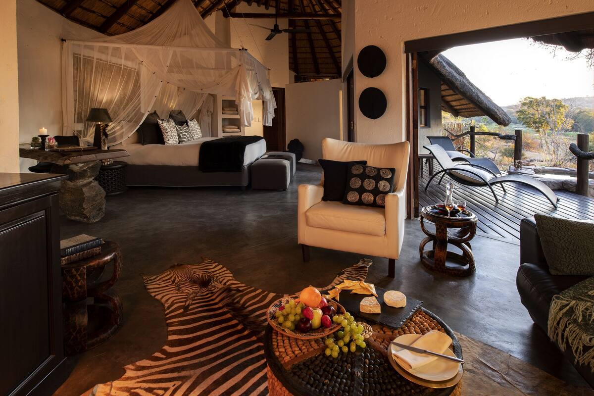 A spacious living area with a bed, lounge chairs, a dining table with fruits and cheese, and a view of the outdoors.