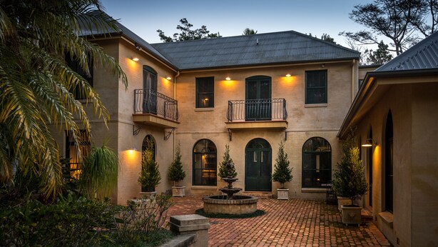 A two-story building with a courtyard, featuring a fountain, potted plants, and a brick pathway.