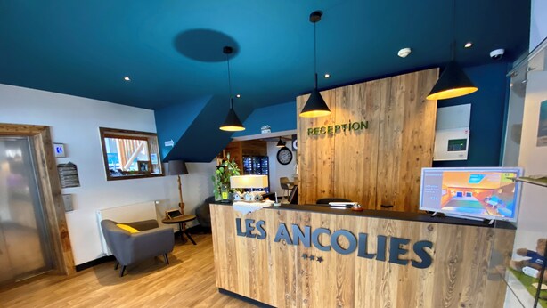 A reception desk with 'LES ANCOLIES' signage, a wooden interior, a reception area with a computer and a TV, and a small seating area with a lamp and a plant. - Hotel Les Ancolies (Beaufort)