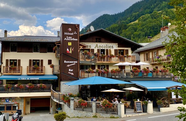 A hotel with a restaurant and bar, outdoor seating with umbrellas, and a mountainous backdrop. - Hotel Les Ancolies (Beaufort)