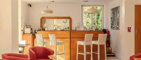A modern kitchen with a wooden counter and white chairs, a dining area with red chairs, and a large window with greenery outside.