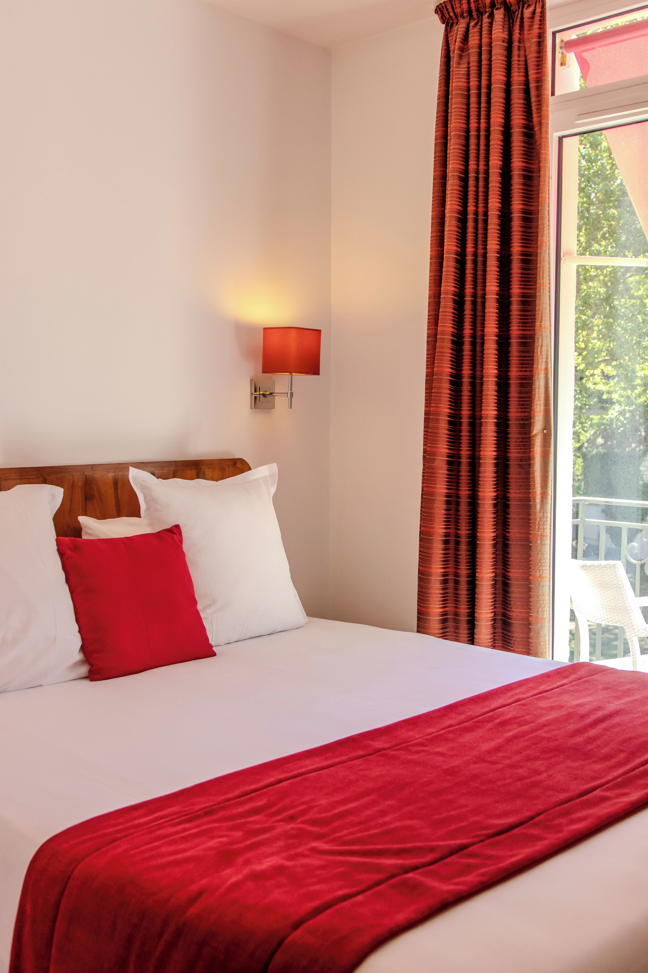 Habitación de hotel con cama, almohadas blancas, una manta roja, una lámpara de pared y un ventanal con cortinas.