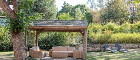 A wooden gazebo with a cushioned bench and a table, surrounded by a stone wall and greenery.