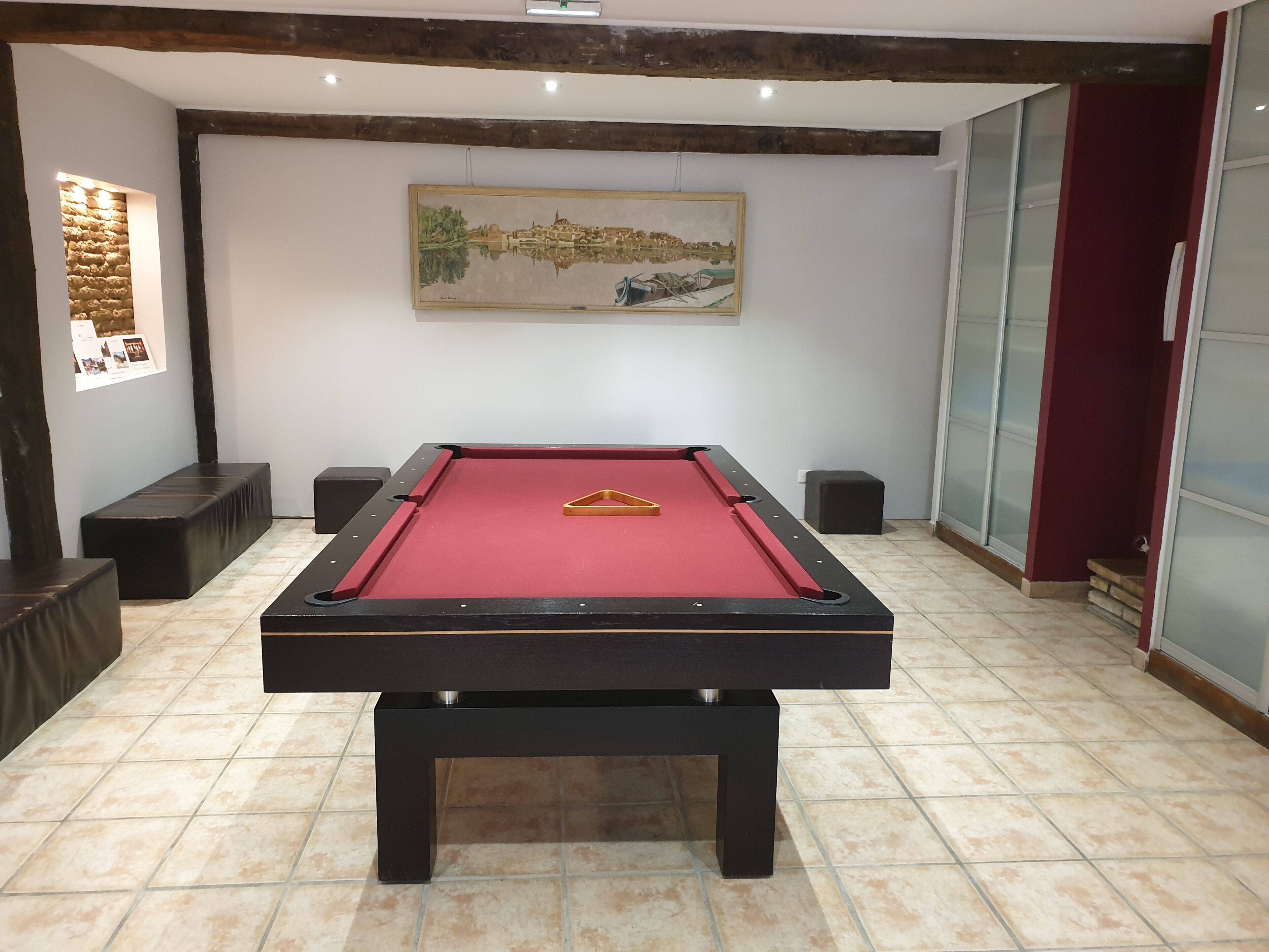 Una mesa de billar con superficie de fieltro rojo, marco negro y una bola de billar amarilla. Hay dos taburetes negros y un cuadro en la pared.
