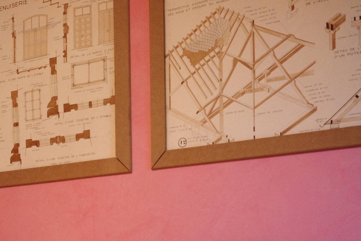 Ein Bett mit weißer Bettwäsche und einem Holz-Kopfende, vor einer rosafarbenen Wand mit zwei gerahmten architektonischen Zeichnungen.