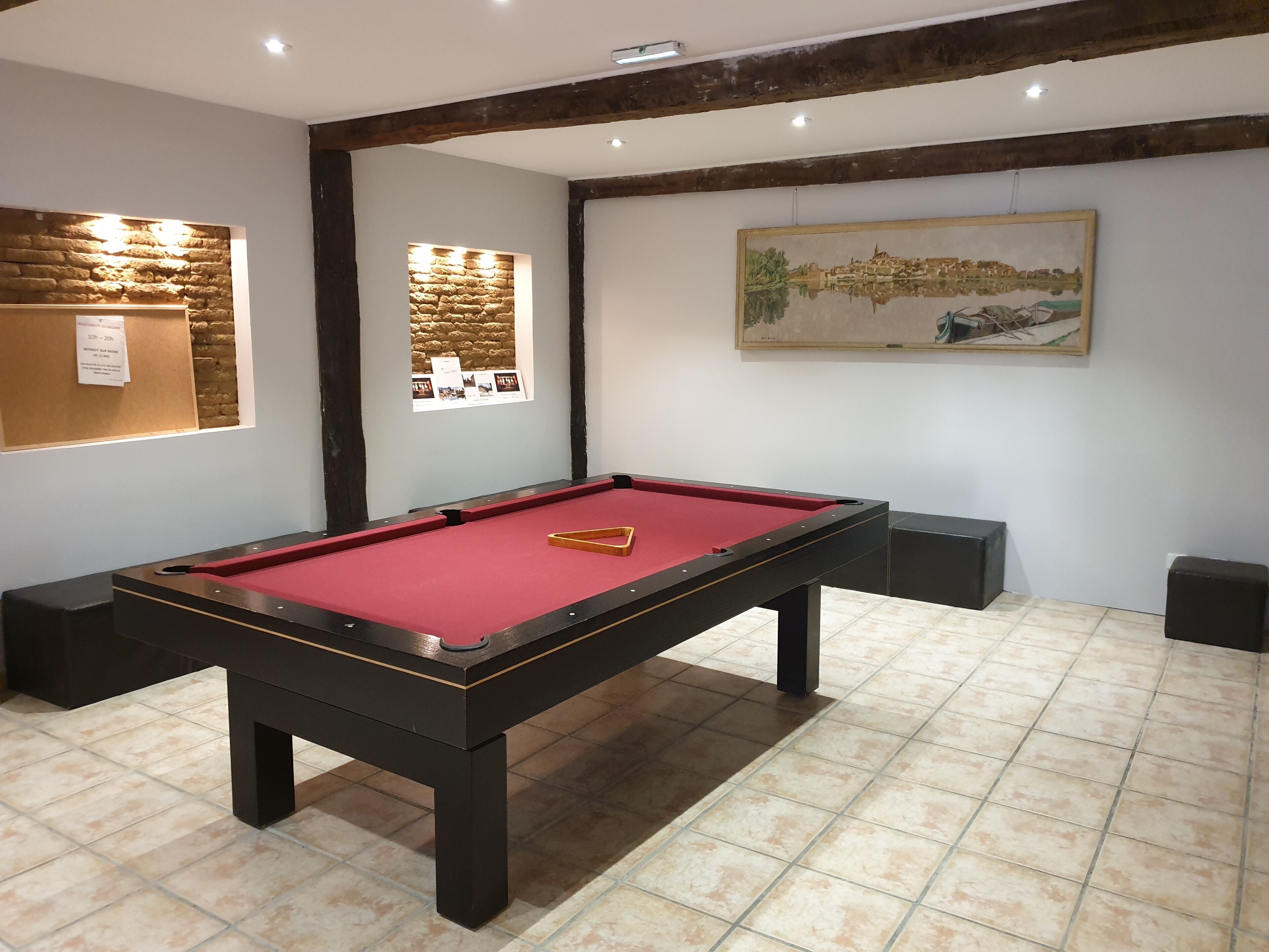 A pool table with a red felt surface, surrounded by dark wooden legs. There is a framed picture on the wall above the table, and a small window with a stone wall design to the left.
