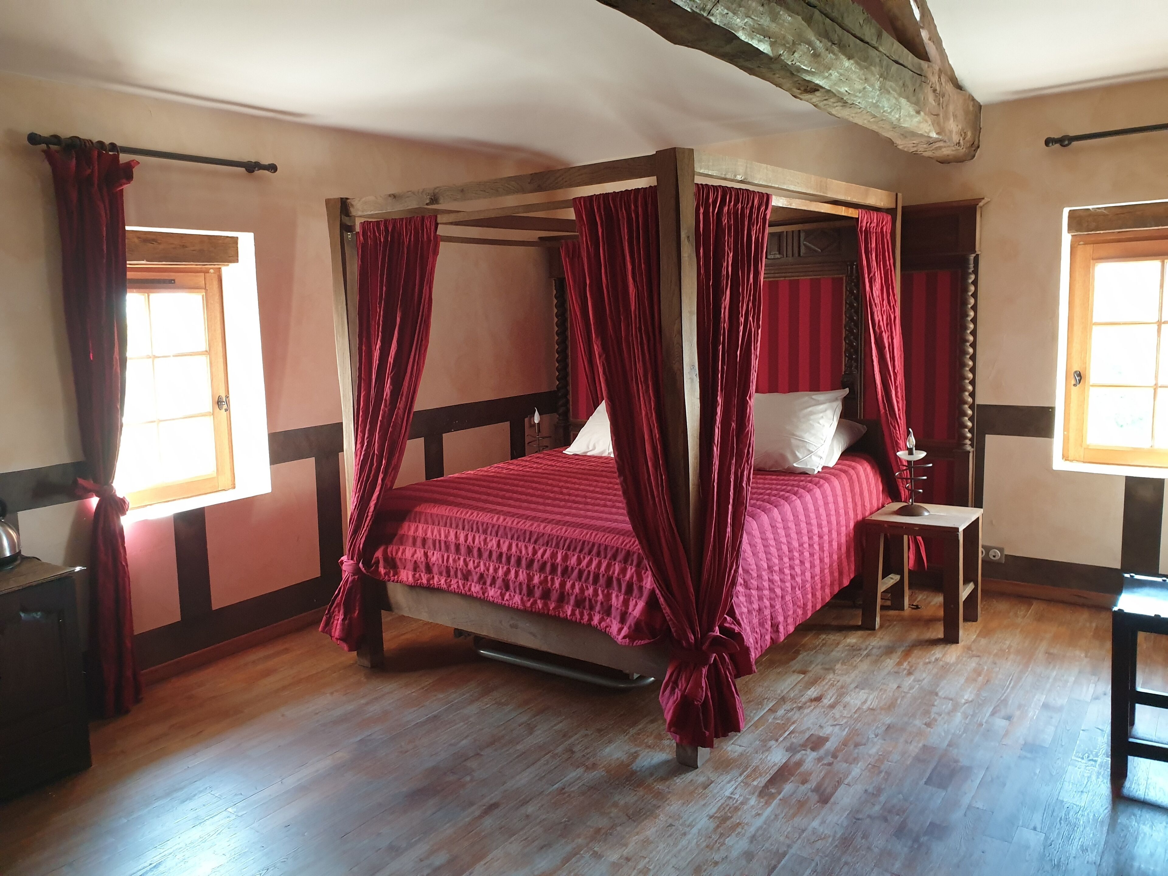 A four-poster canopy bed with red curtains, wooden flooring, and a small bedside table with a lamp.