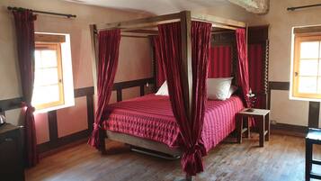 A four-poster canopy bed with red curtains, wooden flooring, and a small bedside table with a lamp.