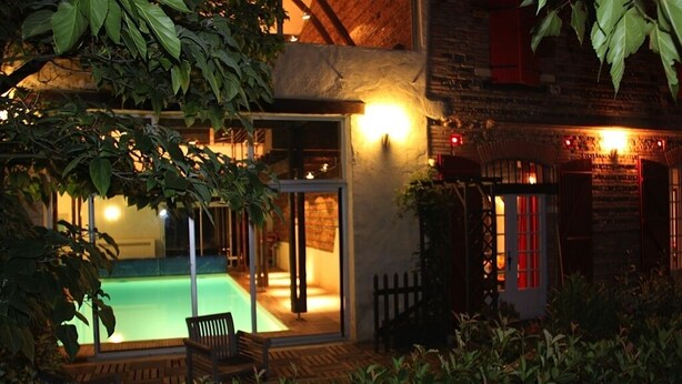 Zona de piscina con cerramiento de vidrio, terraza de madera y un edificio de ladrillo al fondo.