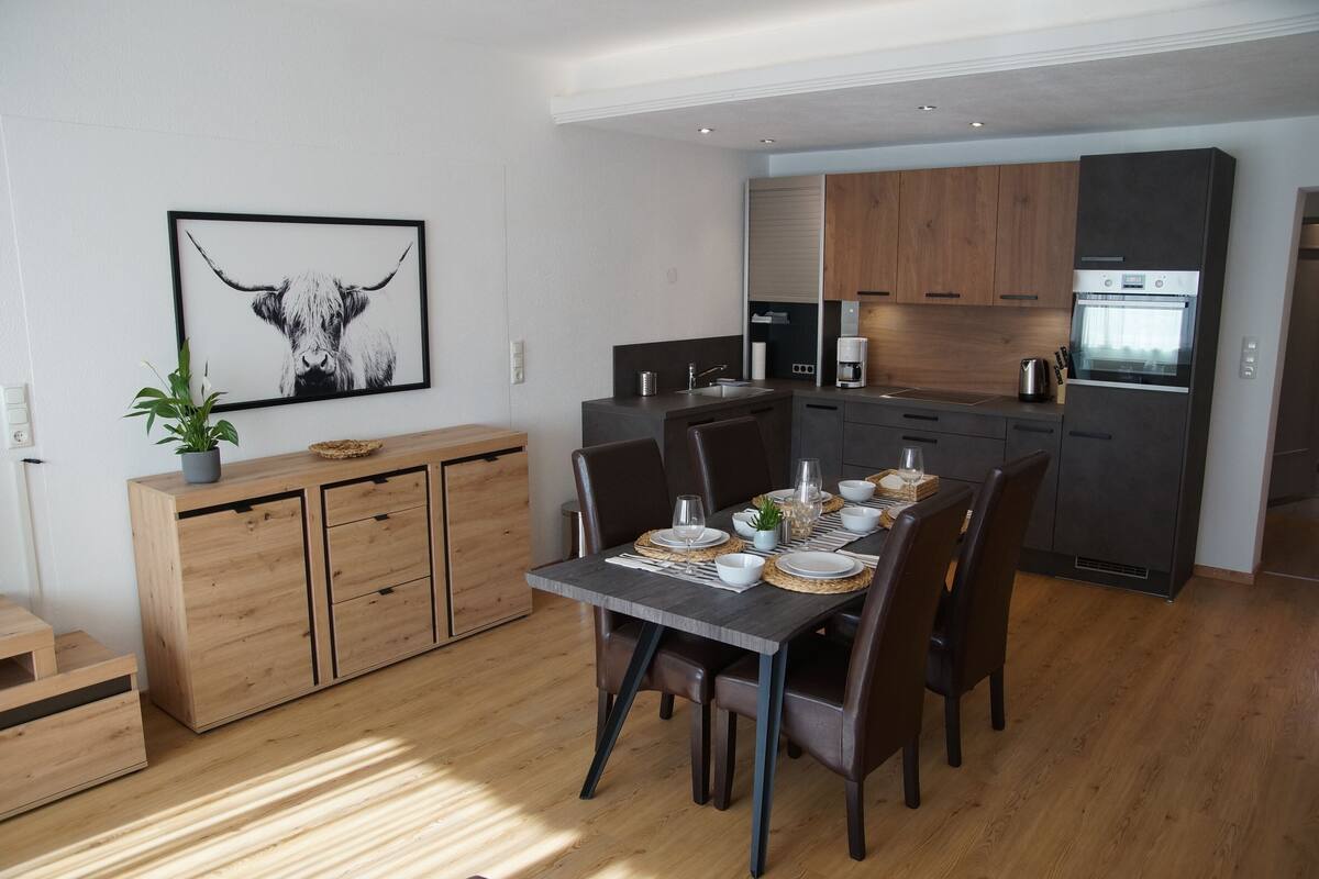 Una cocina moderna con armarios oscuros, horno empotrado y comedor con mesa de madera dispuesta para cuatro comensales.