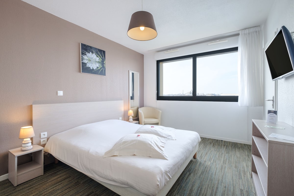 Une chambre d’hôtel avec un grand lit, une table de chevet avec une lampe, une chaise, un bureau avec une télévision et une fenêtre donnant sur un beau paysage.