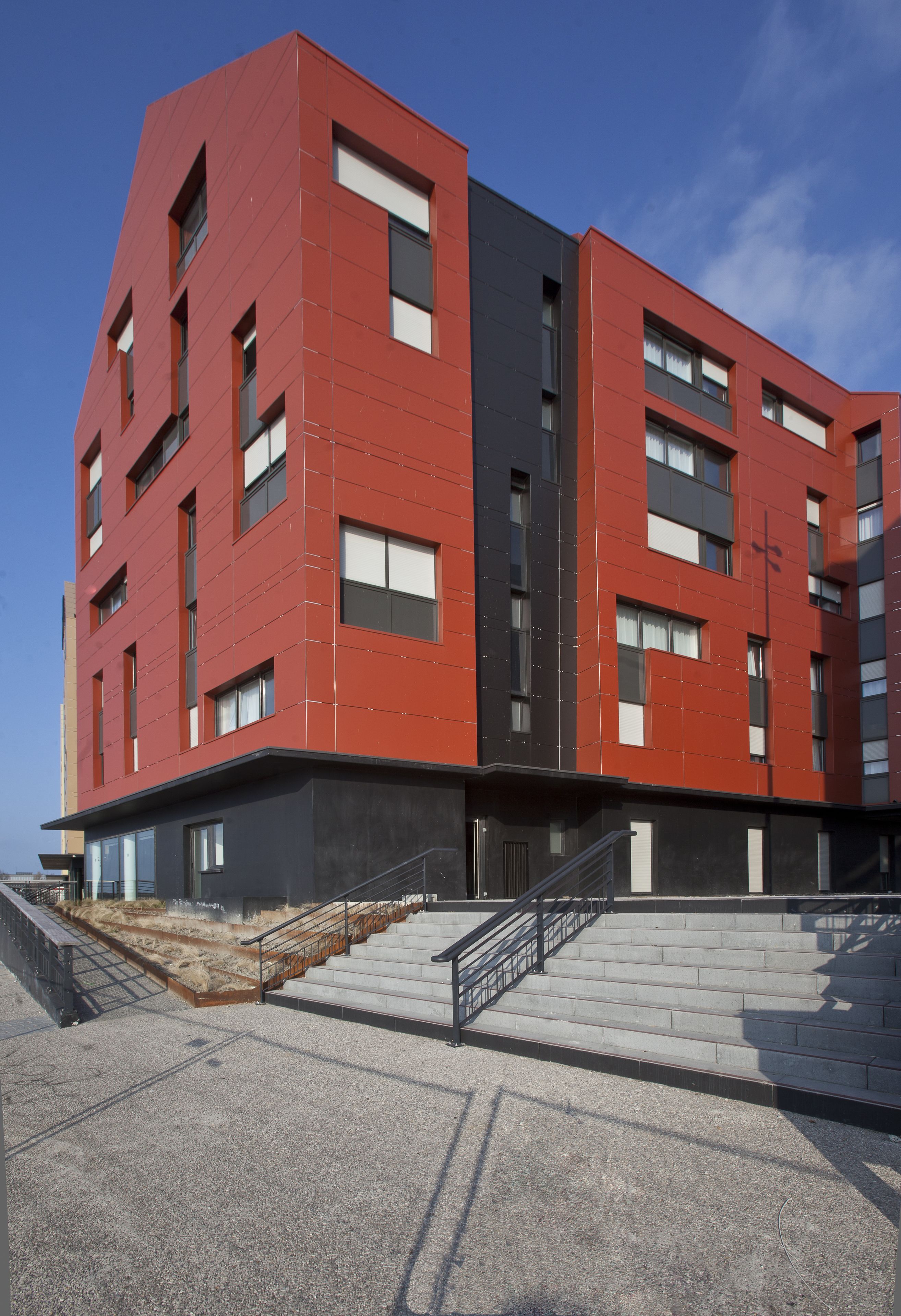 Un bâtiment moderne à la façade rouge et noire, doté de nombreuses fenêtres et d’un escalier menant à l’entrée.