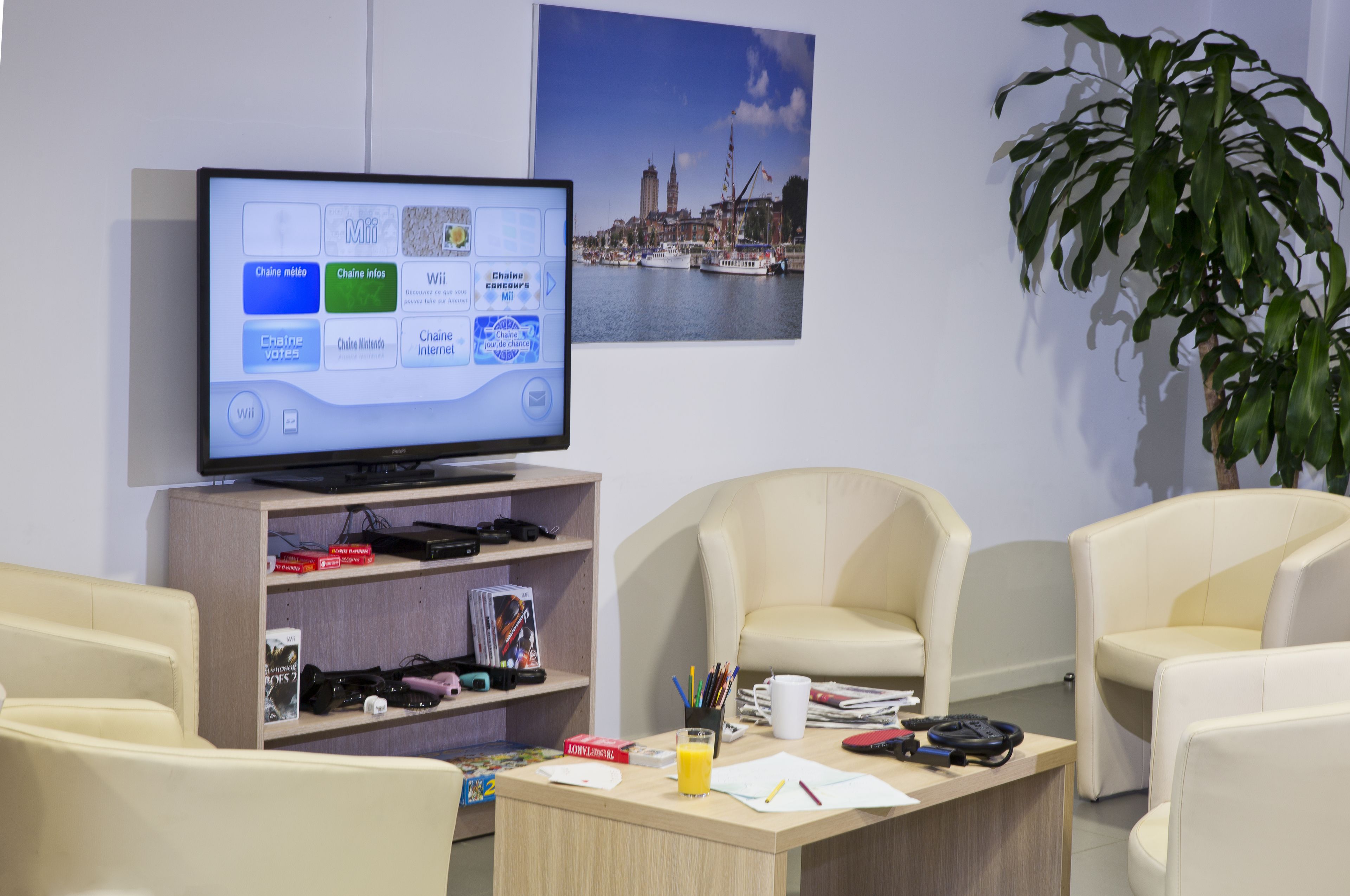 Una habitación de hotel moderna con un televisor de pantalla plana mostrando un menú, una estantería con libros y revistas, una mesa de centro con material de escritorio y un ventanal con vistas a la ciudad.