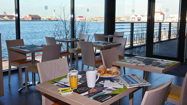 Un comedor con mesas dispuestas para el desayuno, que incluye croissants, un periódico y un vaso de zumo de naranja.