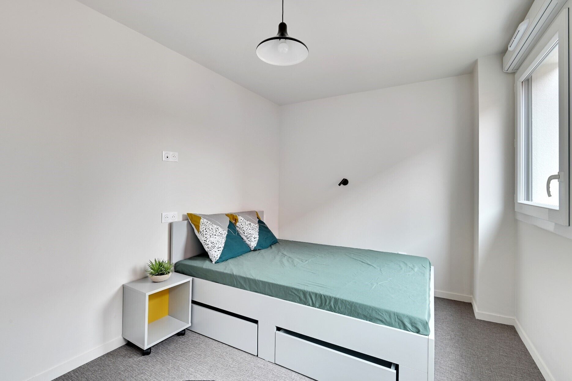 Een kleine, moderne slaapkamer met een enkel bed, een nachtkastje met een plant en een raam met uitzicht.