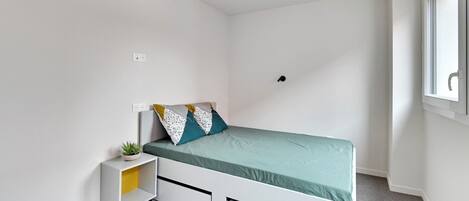 Un dormitorio pequeño y moderno con una cama individual, una mesita de noche con una planta y una ventana con vistas.