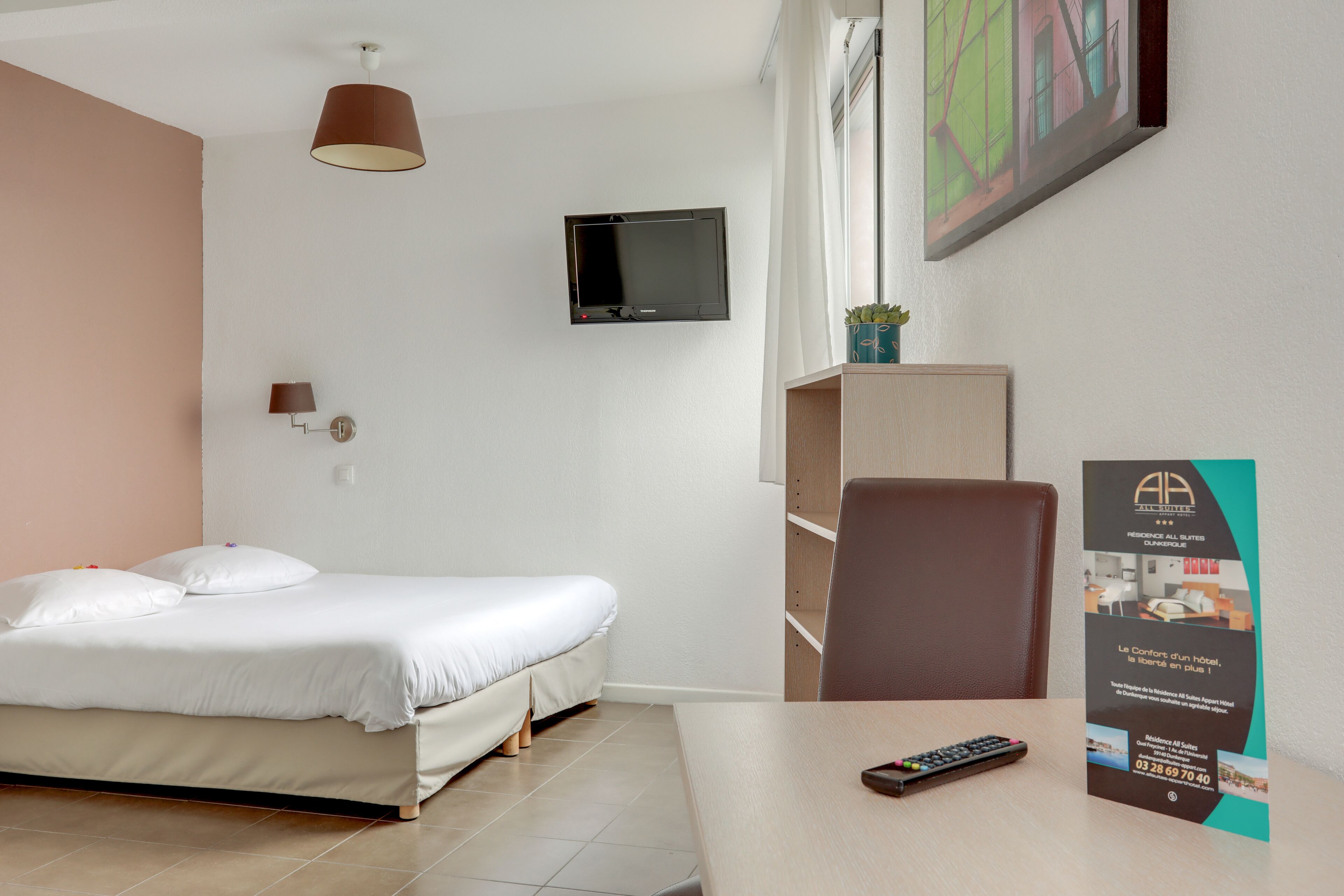 Habitación de hotel con una cama, un televisor montado en la pared, un escritorio con un control remoto y un folleto sobre el escritorio.