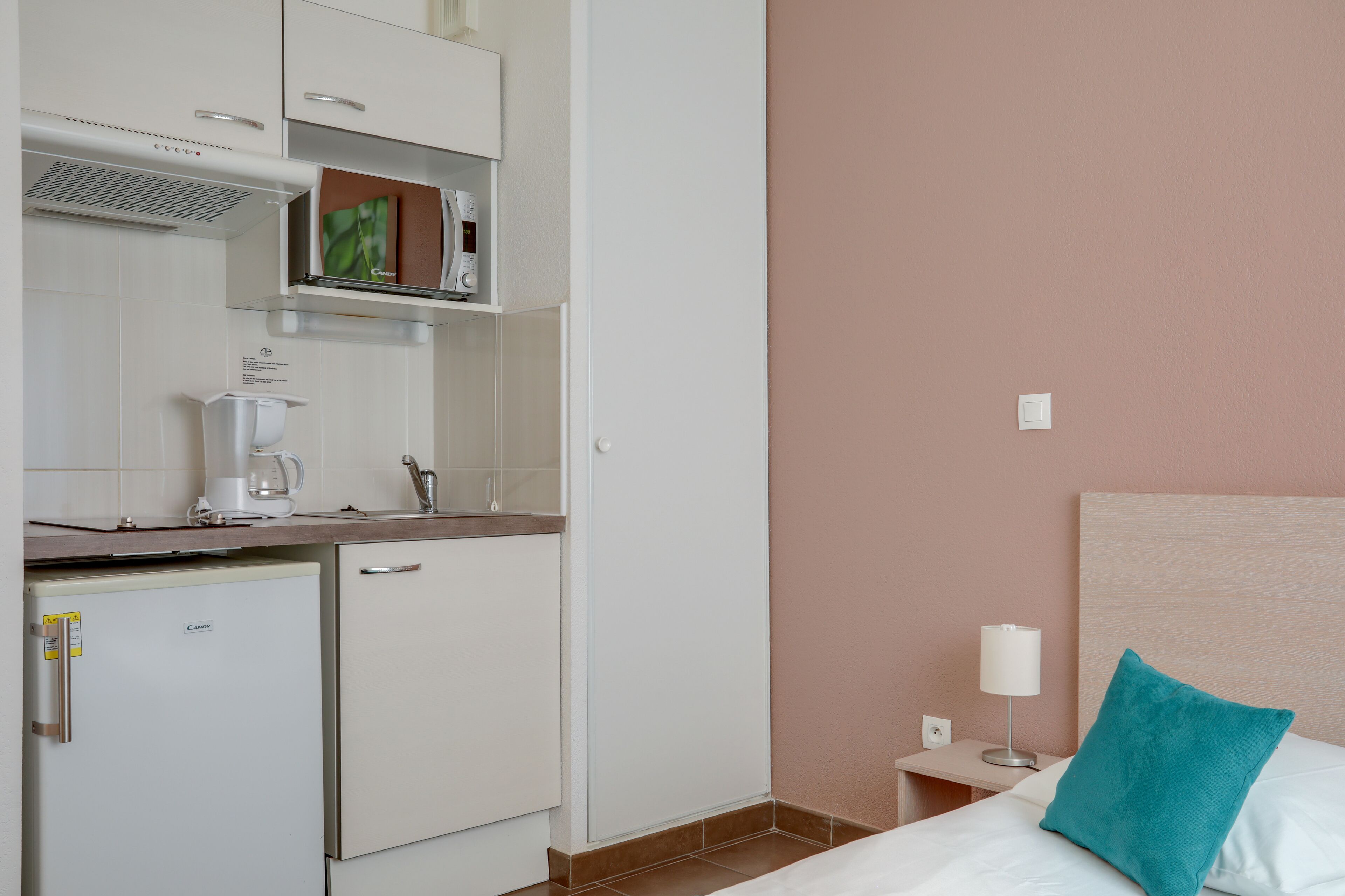 Una camera d'albergo compatta con angolo cottura, un letto con cuscino color petrolio e una lampada da comodino.