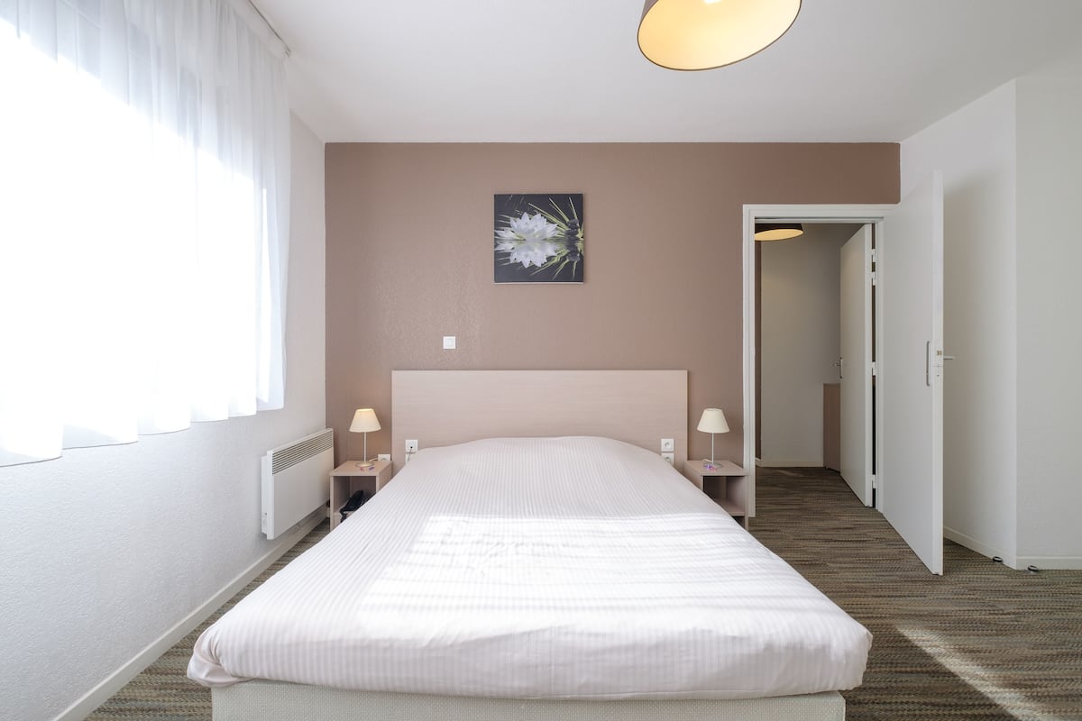 Une chambre d’hôtel avec un grand lit, des lampes de chevet, un radiateur, un tableau au mur et une porte donnant sur une autre pièce.