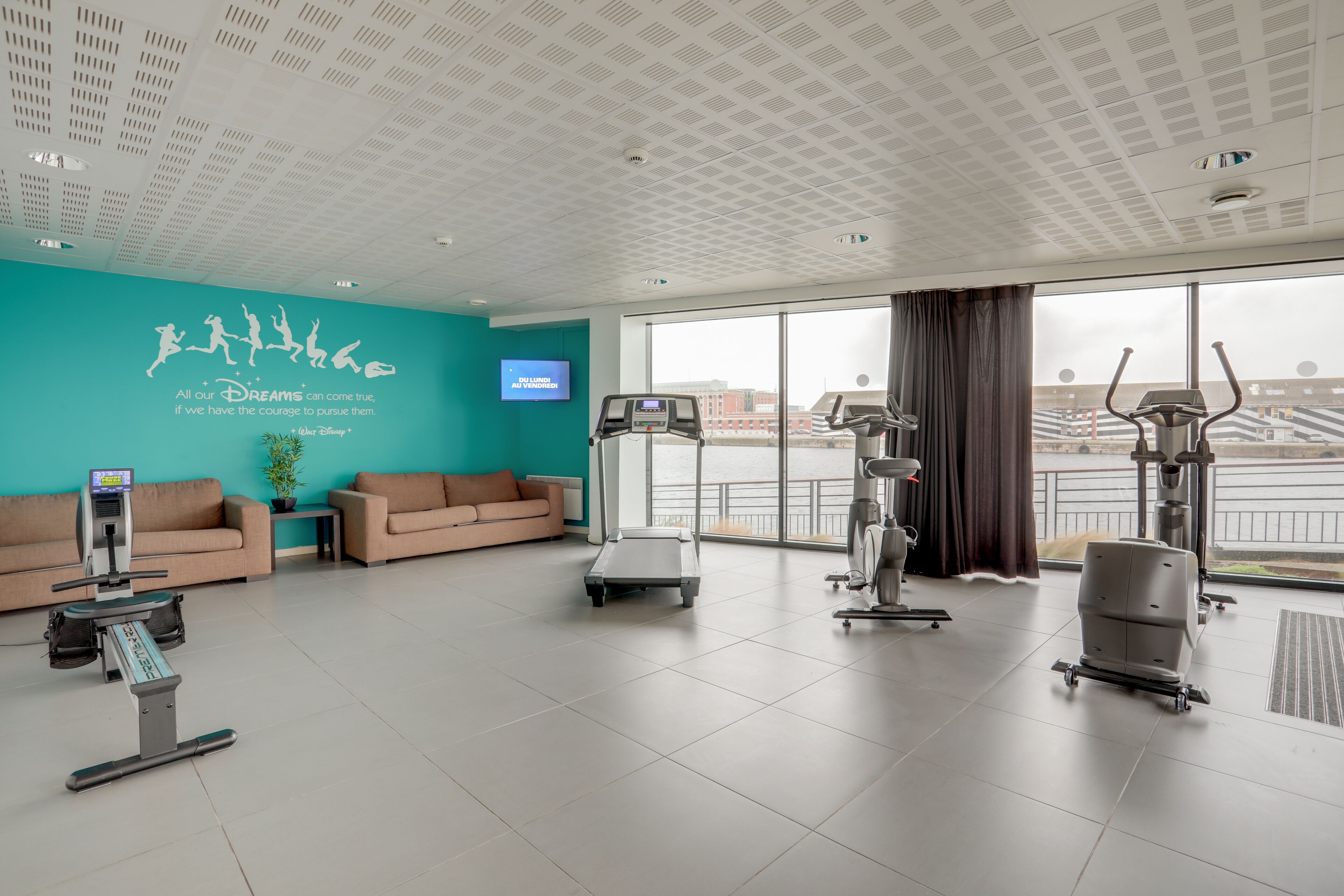 Ein großzügiger Fitnessbereich mit Cardiogeräten, Laufband und Hometrainer. An der Wand ist ein motivierendes Zitat angebracht und ein Fernseher hängt dort.