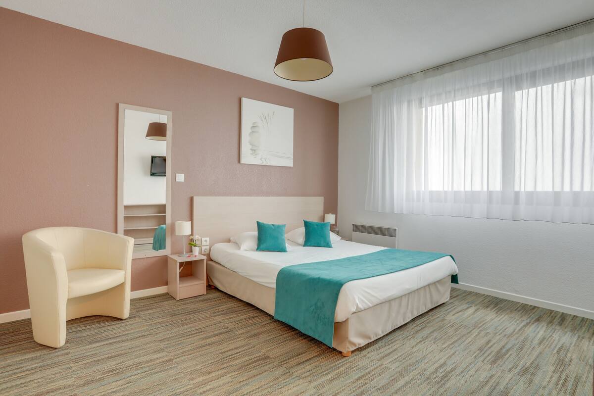Une chambre d’hôtel avec un lit, une chaise, une table de chevet, un miroir et une fenêtre avec des rideaux légers.