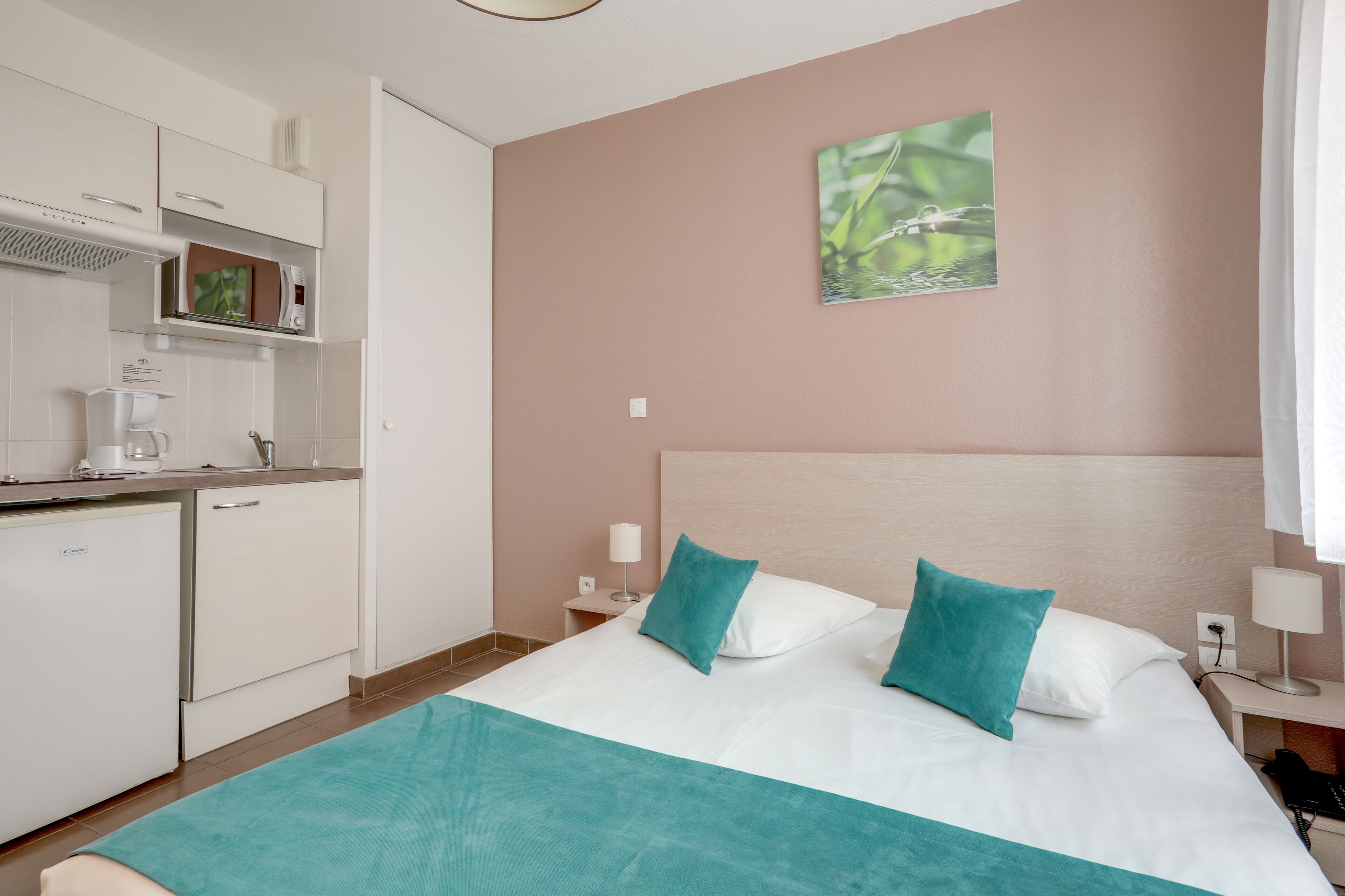 A hotel room with a bed, a desk, a small kitchenette, and a wall-mounted picture.