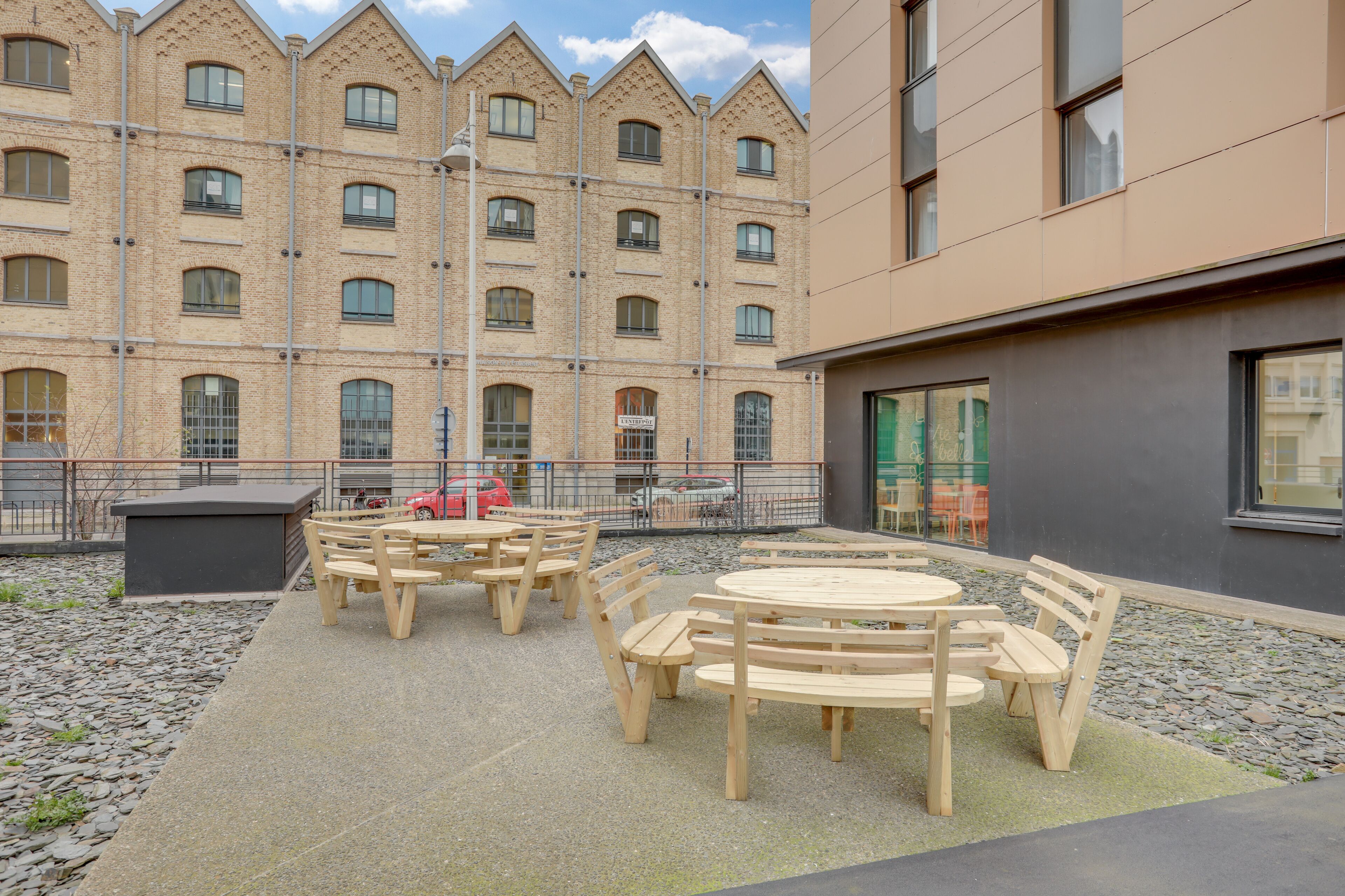 An outdoor seating area with wooden tables and benches, surrounded by a building with a brick facade and multiple windows.