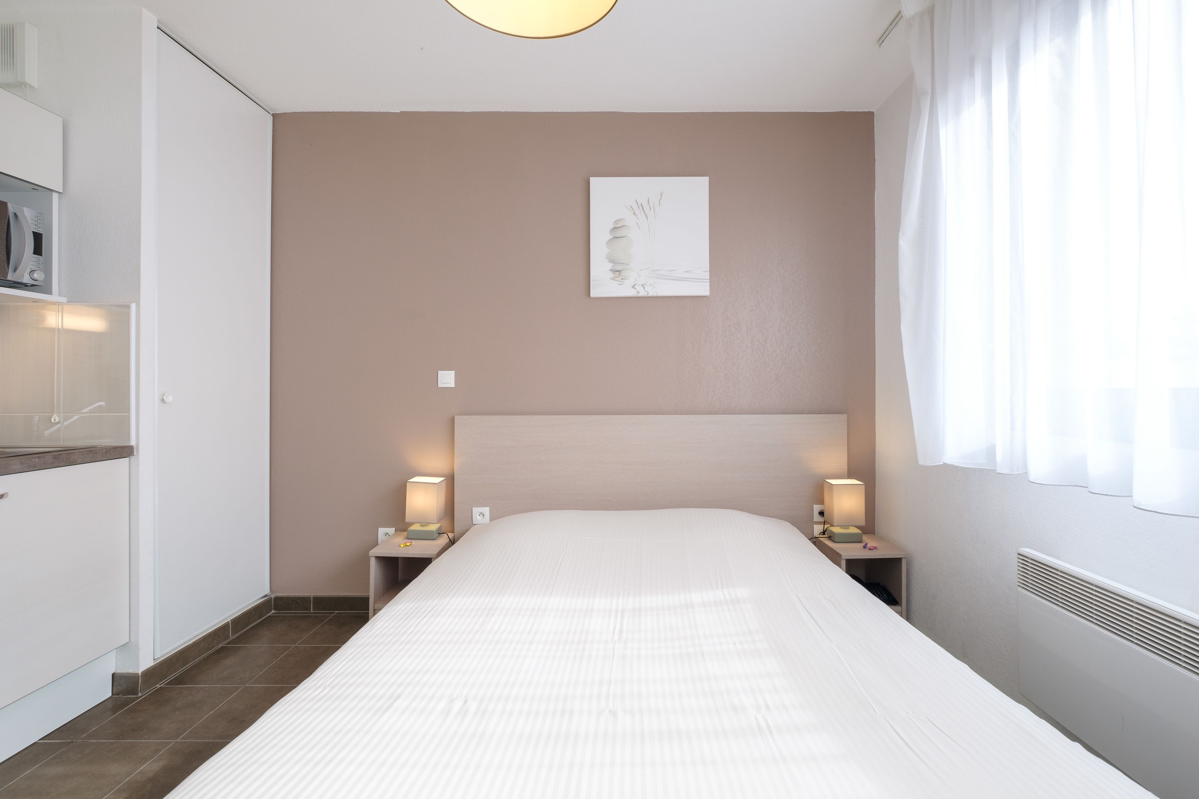 Un dormitorio ordenado con una cama grande, lámparas de noche, una obra de arte en la pared y una ventana con cortinas.