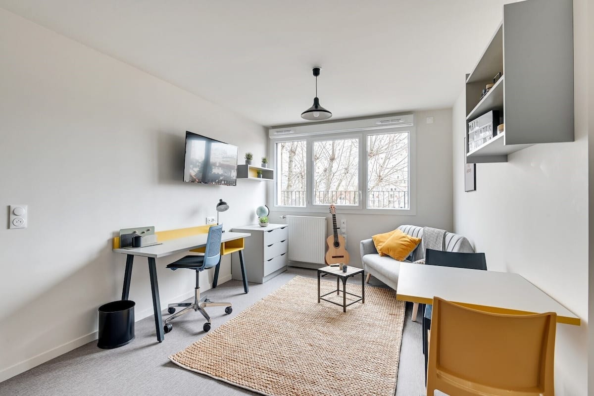 Une pièce compacte et bien éclairée, avec un bureau, une chaise et une petite table. Une guitare est appuyée contre le mur, et une fenêtre laisse entrer la lumière naturelle.