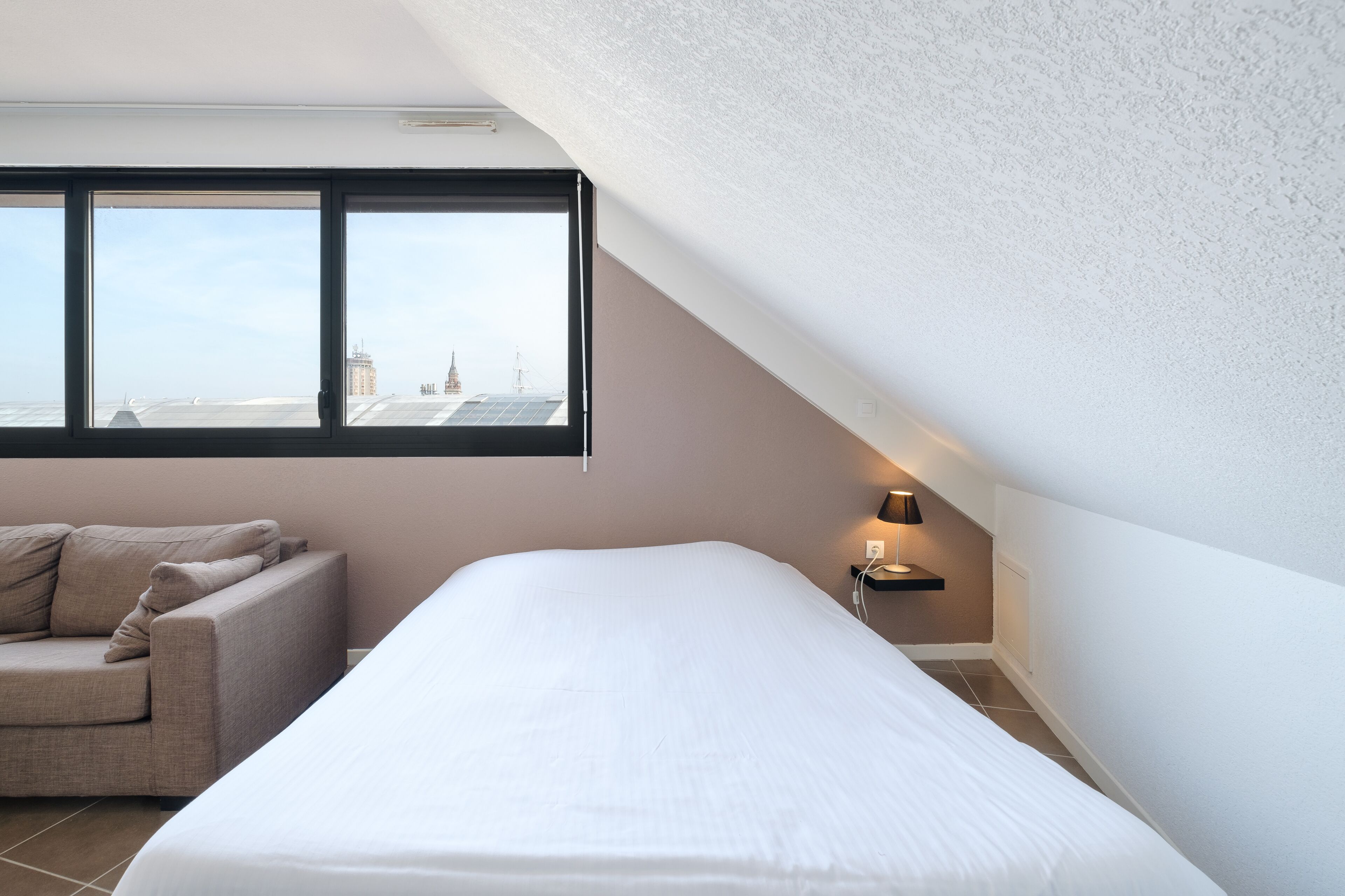 A modern bedroom with a large bed, a sofa, and a window offering a city view.