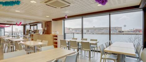 A modern dining area with tables and chairs, large windows with a view of a river and cityscape, and ceiling decorations.