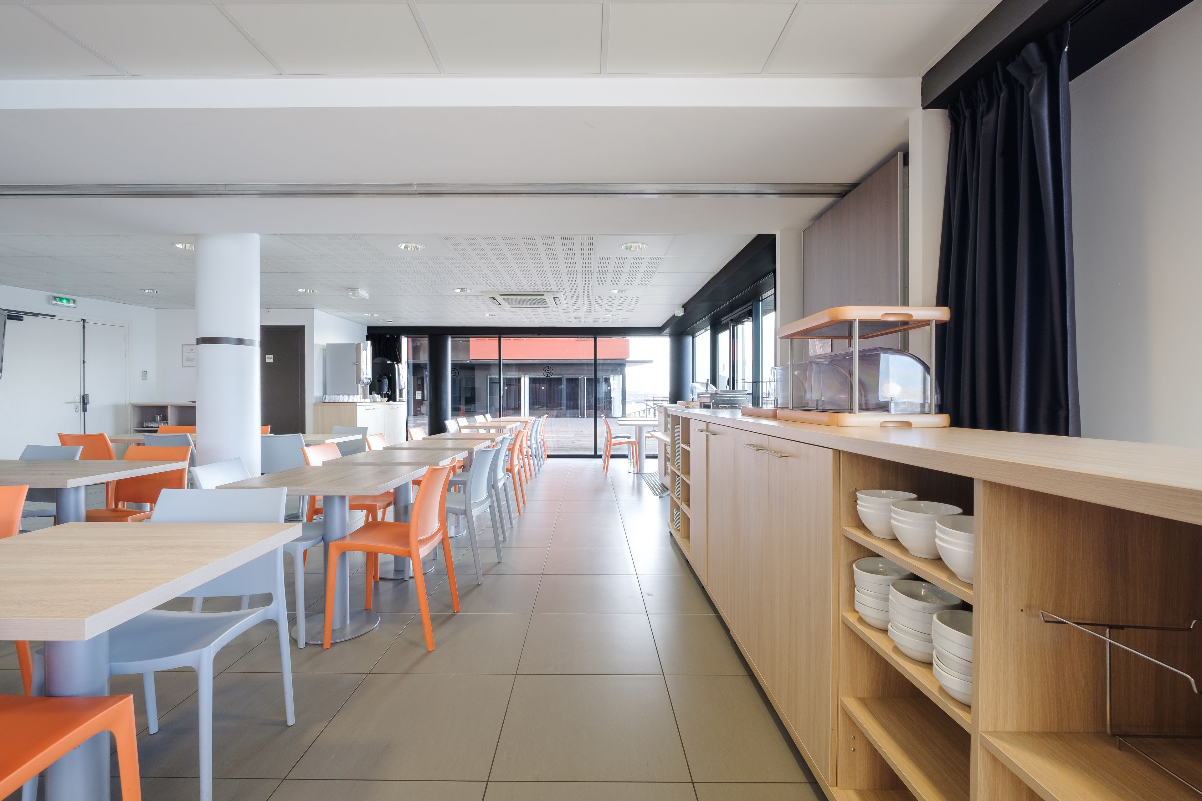 Une cafétéria moderne avec de longues tables et des chaises, un comptoir où sont disposés des bols, et une vue sur l’extérieur.