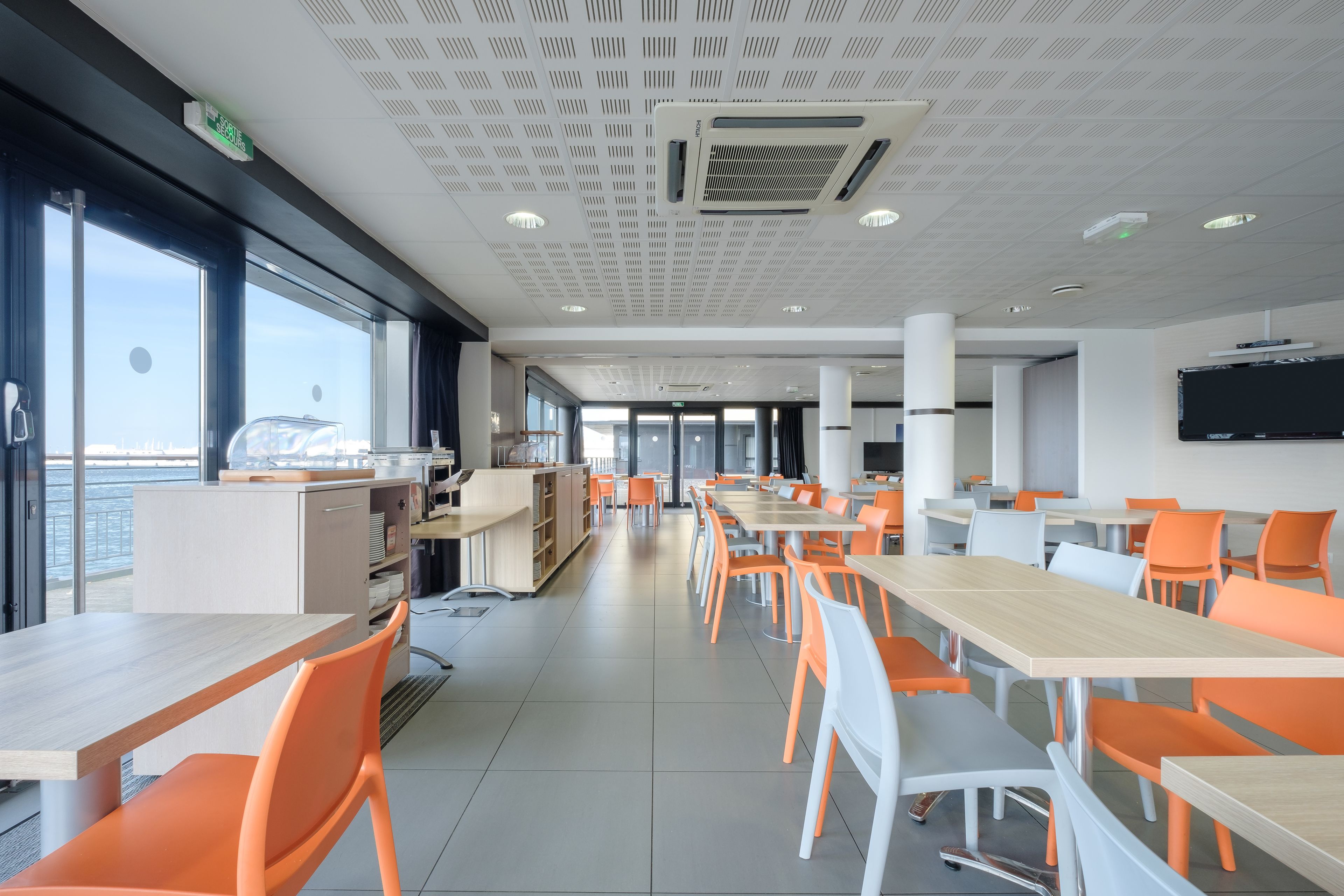 Eine moderne Cafeteria mit orangefarbenen und weißen Stühlen, großen Tischen und Meerblick durch die Fenster.