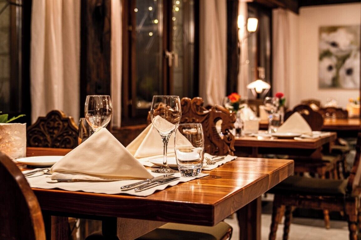 A dining table set with white napkins, wine glasses, and water glasses, with a wooden chair and a decorative vase in the background.
