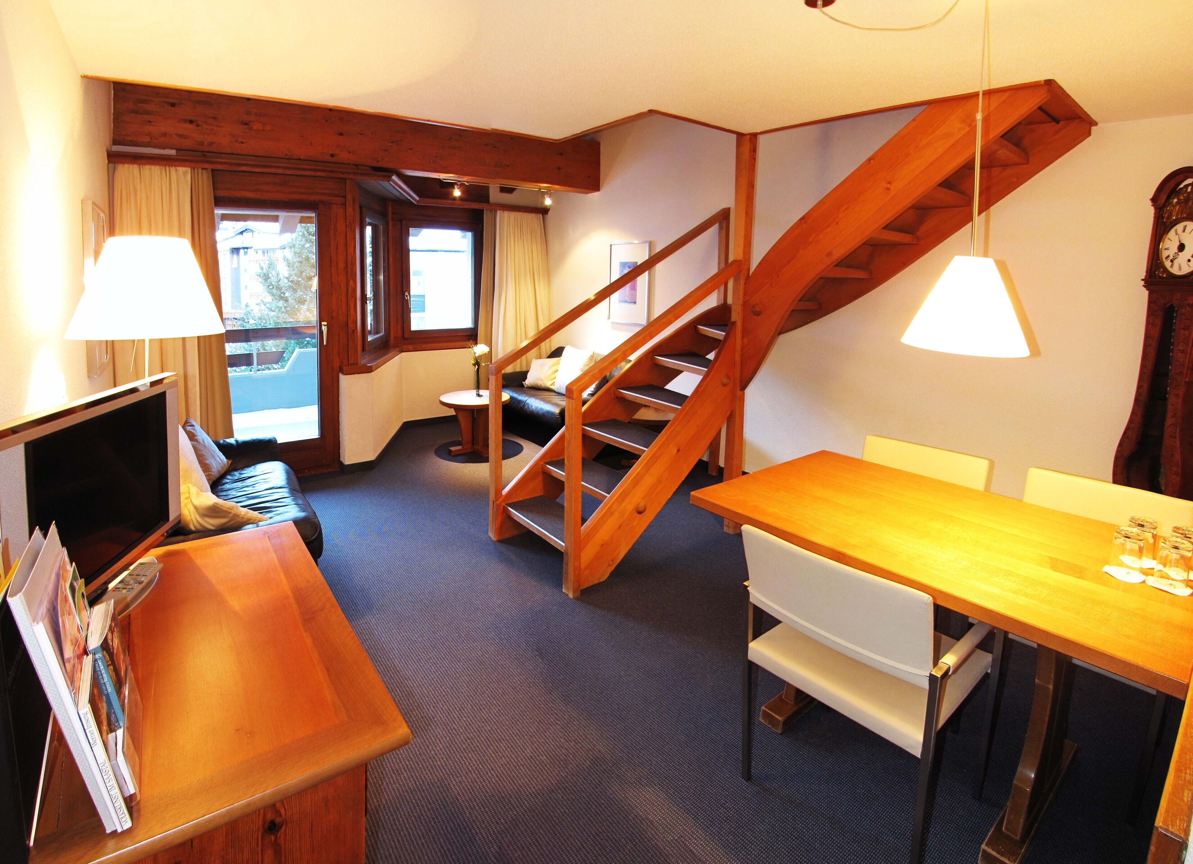 A room with a wooden staircase, a dining table, a sofa, and a TV.