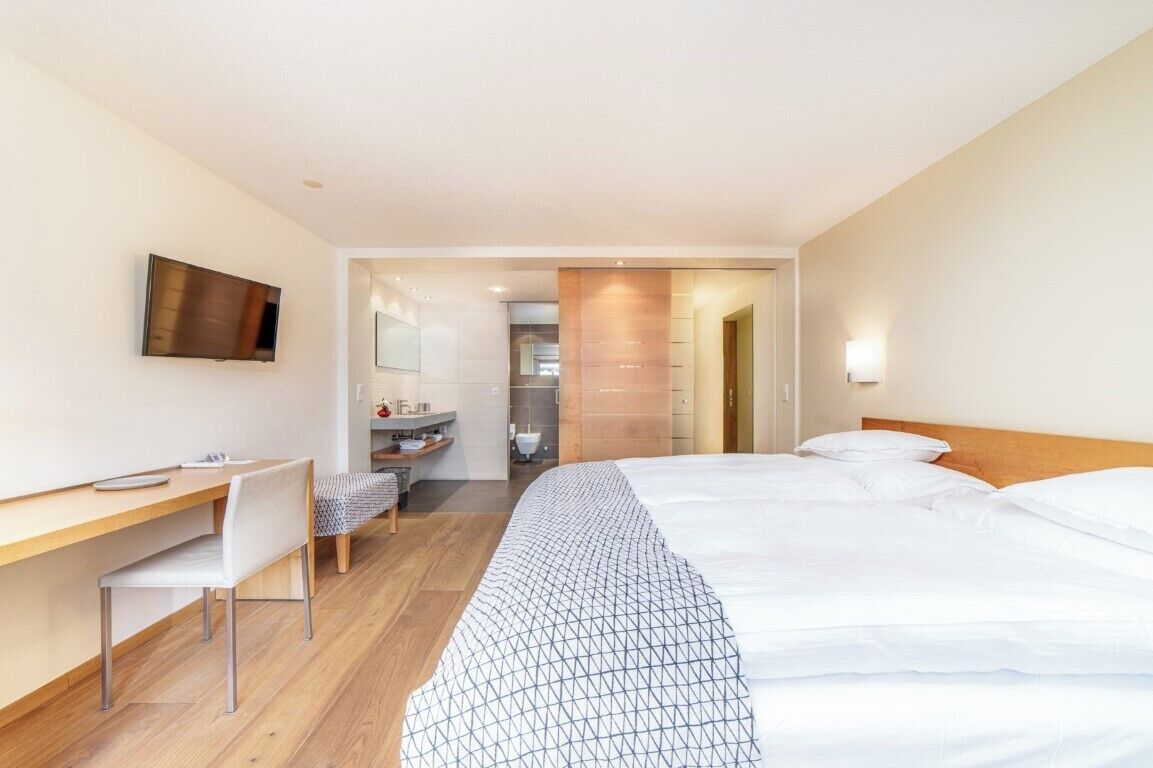 Une chambre d’hôtel moderne équipée d’un lit, d’un bureau, d’une chaise, d’une télévision et d’une salle de bain visible à travers une porte ouverte.