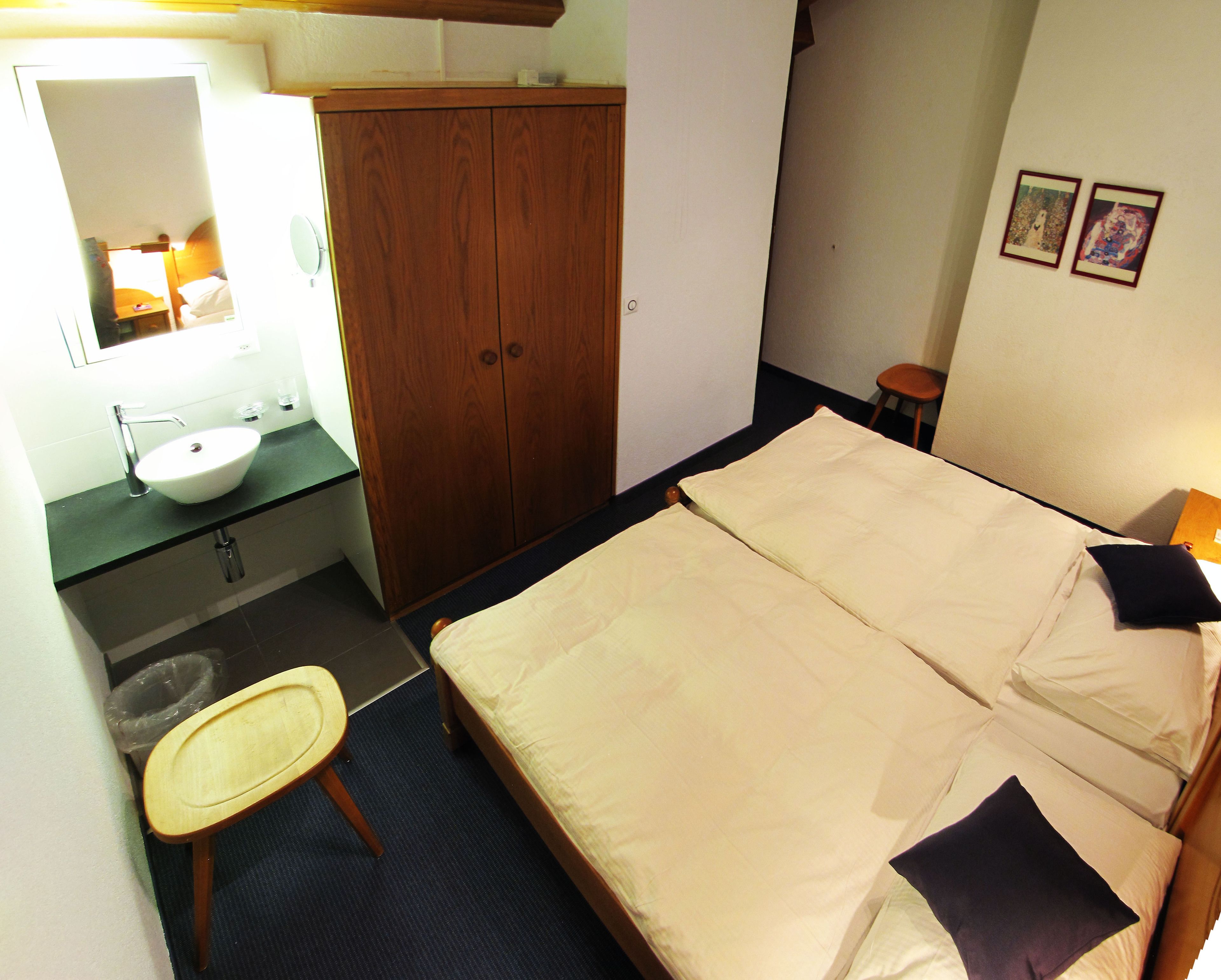 A bedroom with a bed, a wooden wardrobe, a small round table, a sink, and a mirror.