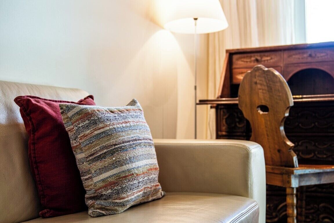 Un canapé beige agrémenté d’un coussin à rayures rouges et multicolores, une chaise en bois et une lampe dans une pièce où se trouve un meuble en bois.