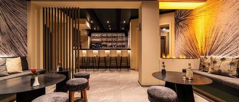 Un bar moderno con barra, taburetes y una zona de estar con sofá y mesas redondas.