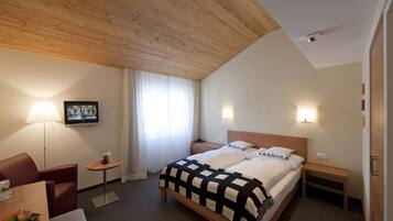 Habitación de hotel con techo de madera, cama con colcha a cuadros blanco y negro, televisor en la pared, escritorio con lámpara y una mesita con fruta.