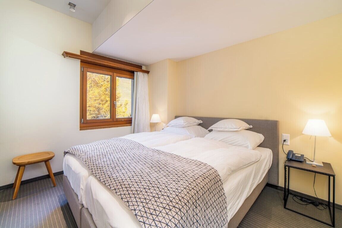 Een hotelkamer met een bed, twee bedlampen, een nachtkastje, een raam met gordijnen, en een klein houten krukje.