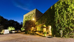 Un edifici amb un carrer de grava, cotxes estacionats, mobiliari exterior i una entrada ben il·luminada.
