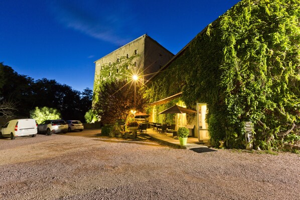 A building with a gravel driveway, parked cars, outdoor seating, and a well-lit entrance.