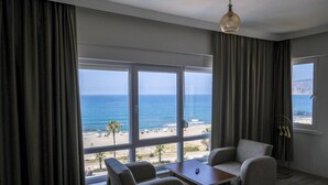 A room with a large window overlooking a beach, a sofa, a table, and a lamp. - Hotel Bella Bravo (Alanya)