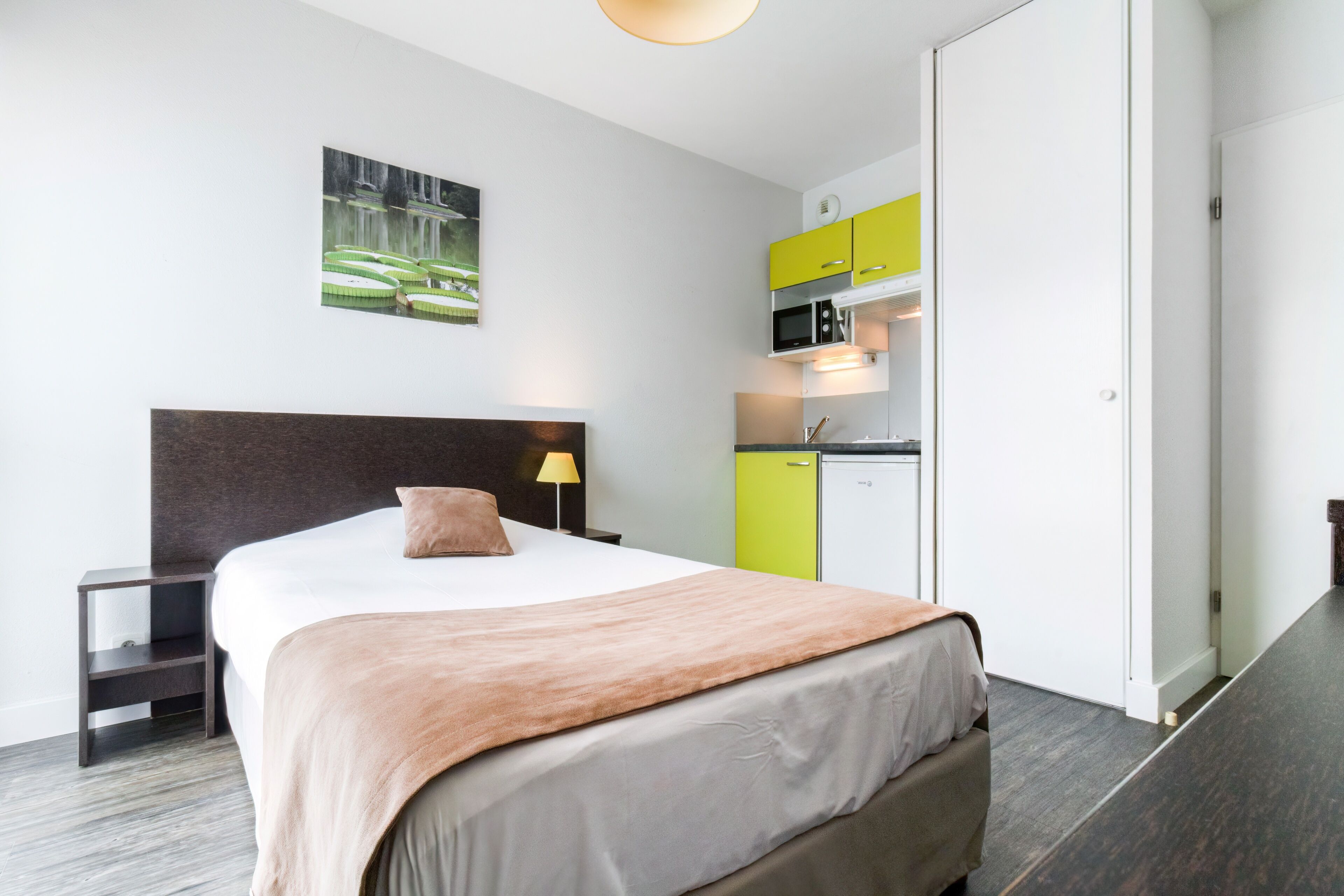 A modern hotel room with a large bed, a bedside table, a lamp, a painting on the wall, and a kitchenette with yellow cabinets.