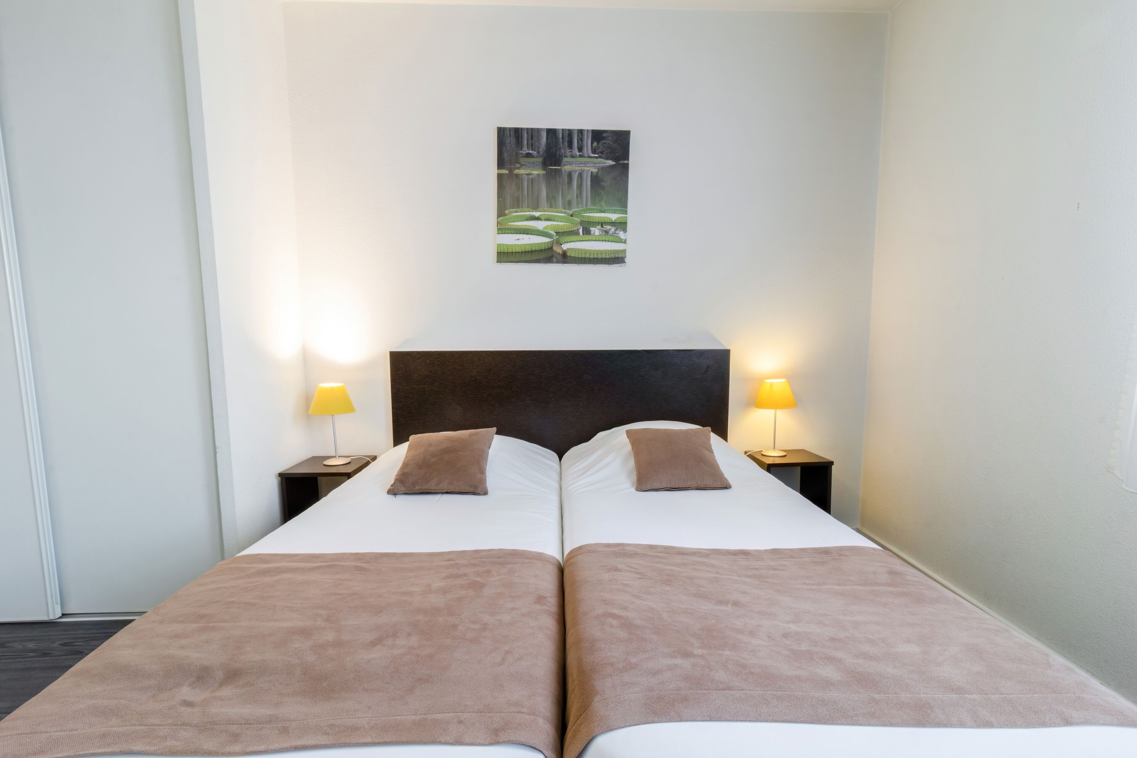 A double bed with two pillows and a headboard, flanked by two bedside tables with lamps, in a room with a painting on the wall.