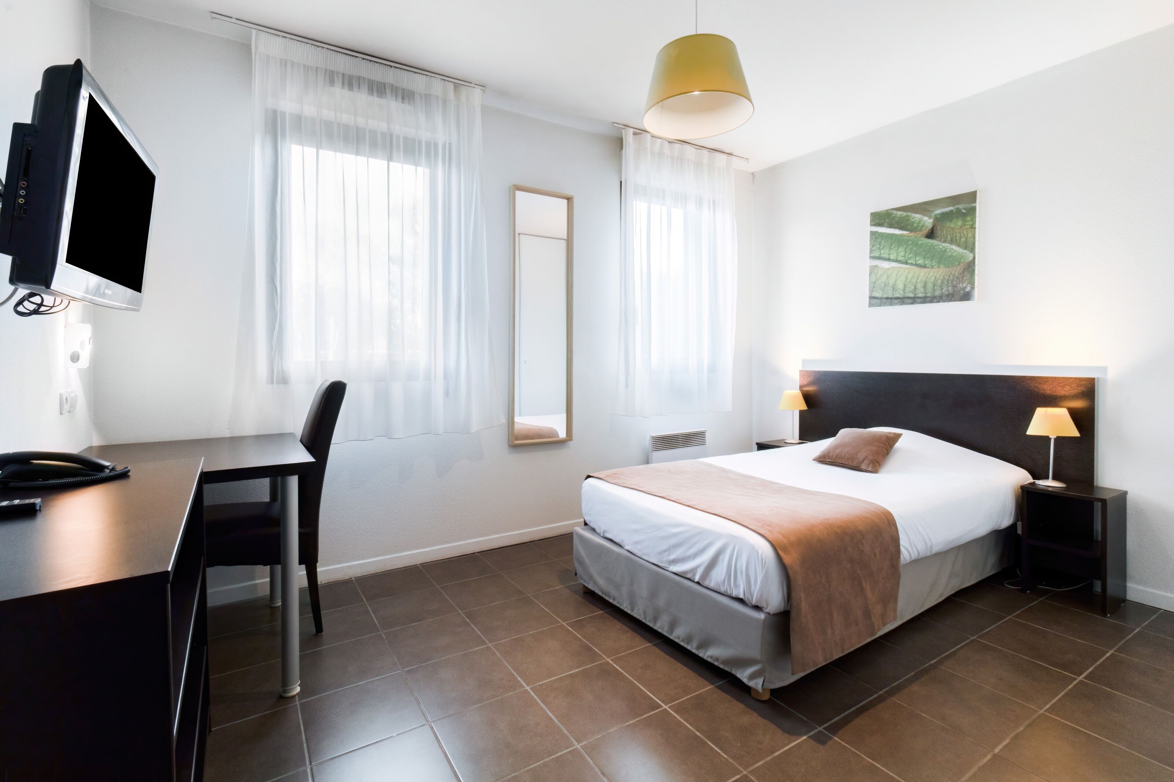 Une chambre d’hôtel moderne équipée d’un lit, de lampes de chevet, d’un bureau avec une chaise, d’une télévision et d’une grande fenêtre avec des rideaux légers.
