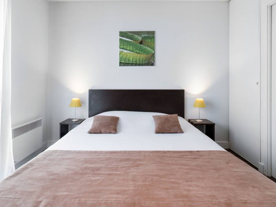 A neatly made bed with a brown blanket and two pillows, a black headboard, and a wall-mounted picture of a green landscape.